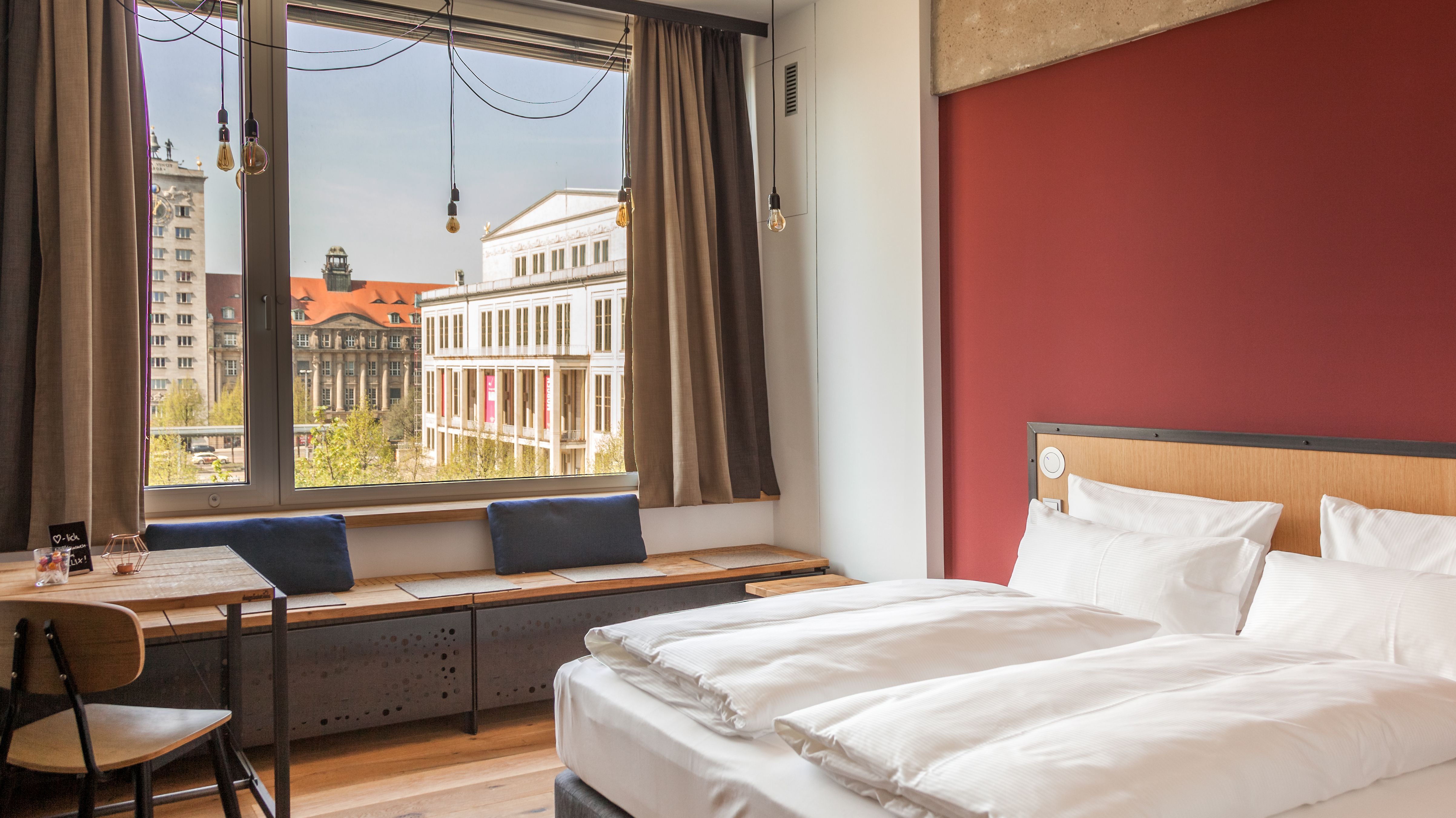Ein Hotelzimmer mit einem Doppelbett mit Blick auf die Oper Leipzig. Im Zimmer ist noch ein Tisch mit einem Stuhl, vor dem Fenster ist eine Sitzbank aus Holz mit zwei Kissen. 