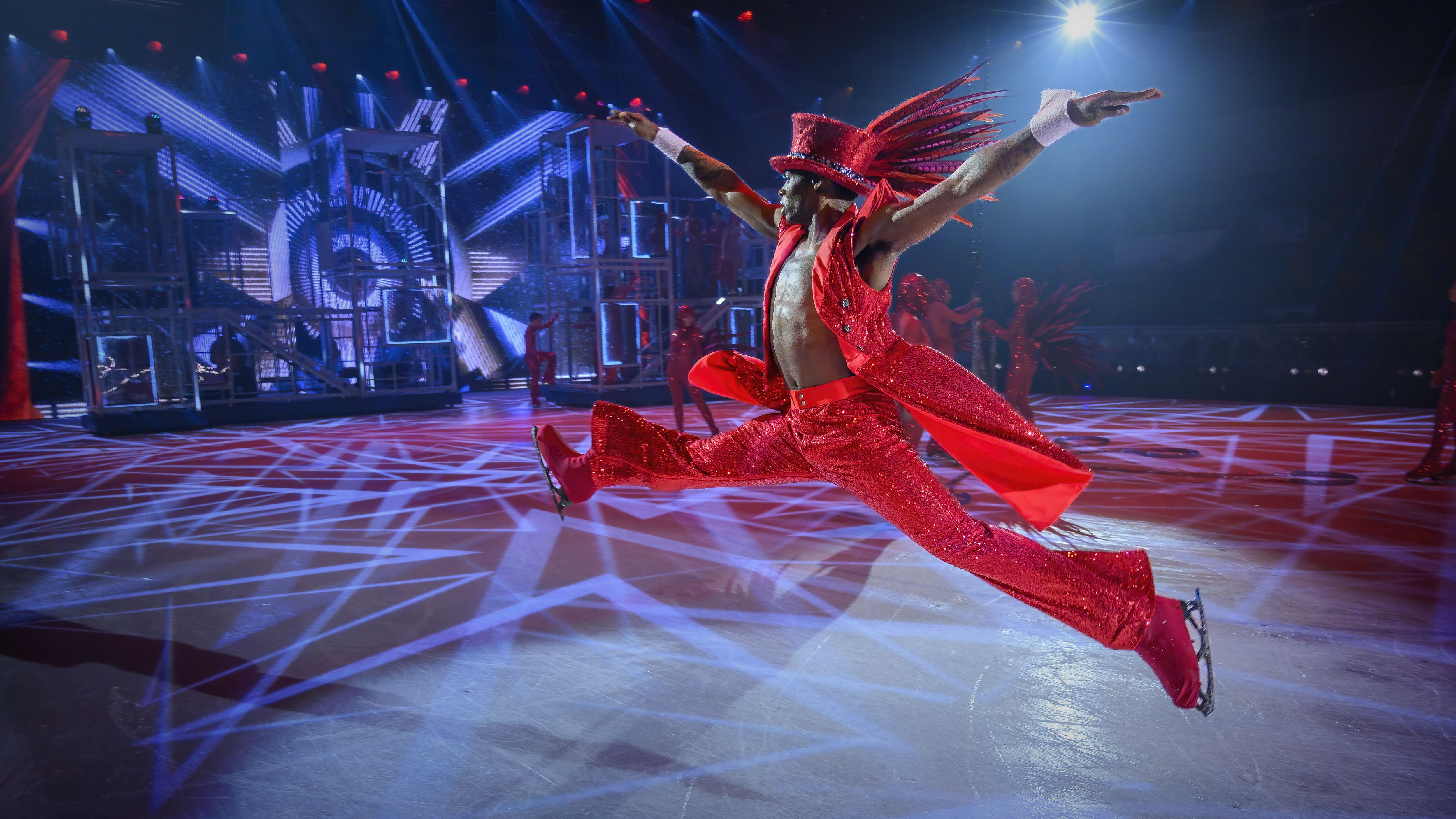 Ein Eiskunstläufer in einem roten, glitzernden Anzug macht einen Sprung mit weit gespreizten Beinen.  