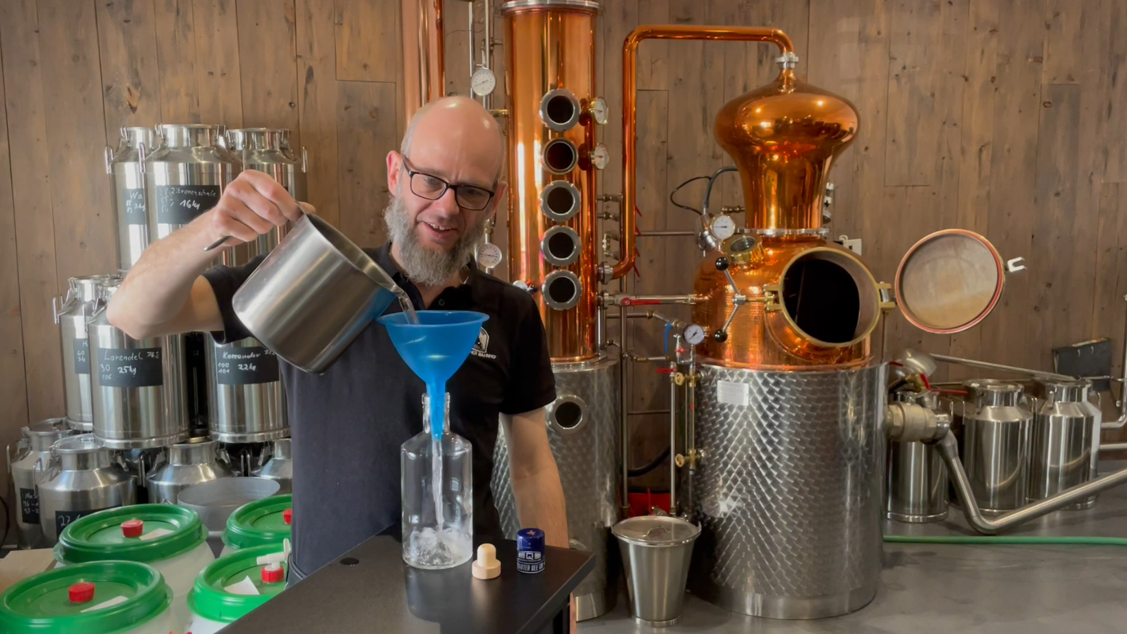 Ein lächelnder Mann mit Brille und Bart füllt in einer Destillerie Flüssigkeit aus einem Metallgefäß durch einen blauen Trichter in eine Glasflasche.