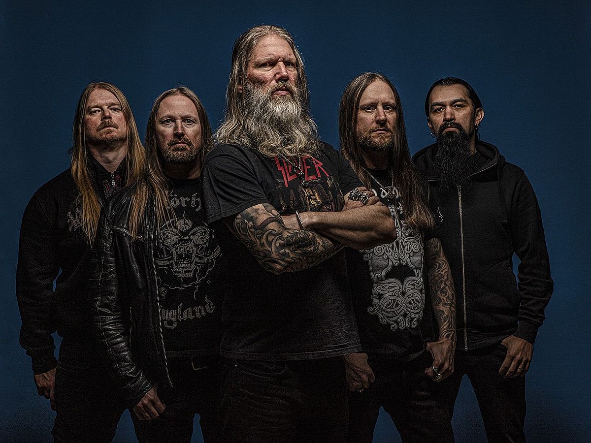 Fünf Männer stehen in einer Dreiecksform vor einer dunkelblauen Wand. Alle tragen schwarze Kleidung und haben einen Bart und lange Haare. 