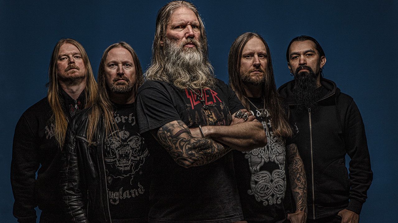 Fünf Männer stehen in einer Dreiecksform vor einer dunkelblauen Wand. Alle tragen schwarze Kleidung und haben einen Bart und lange Haare. 