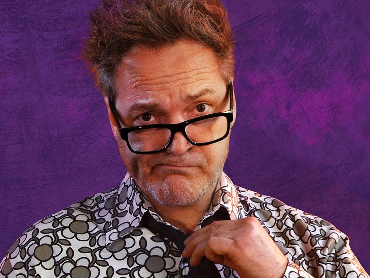 Paul Pnazer steht vor einem lila Hintergrund. Er trägt ein weiß-schwarz-grün-meliertes Hemd und eine schwarze Krawatte. 