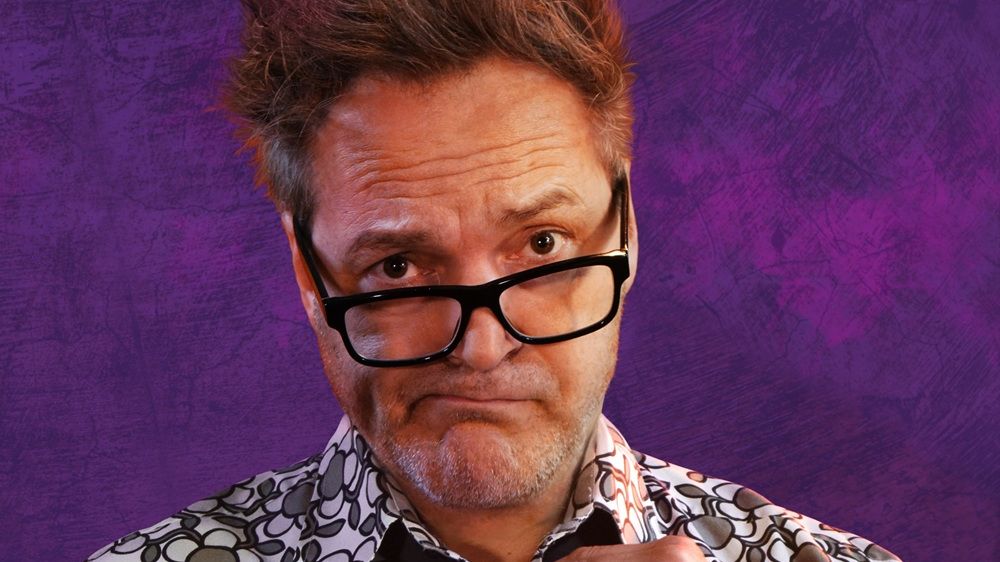 Paul Pnazer steht vor einem lila Hintergrund. Er trägt ein weiß-schwarz-grün-meliertes Hemd und eine schwarze Krawatte. 