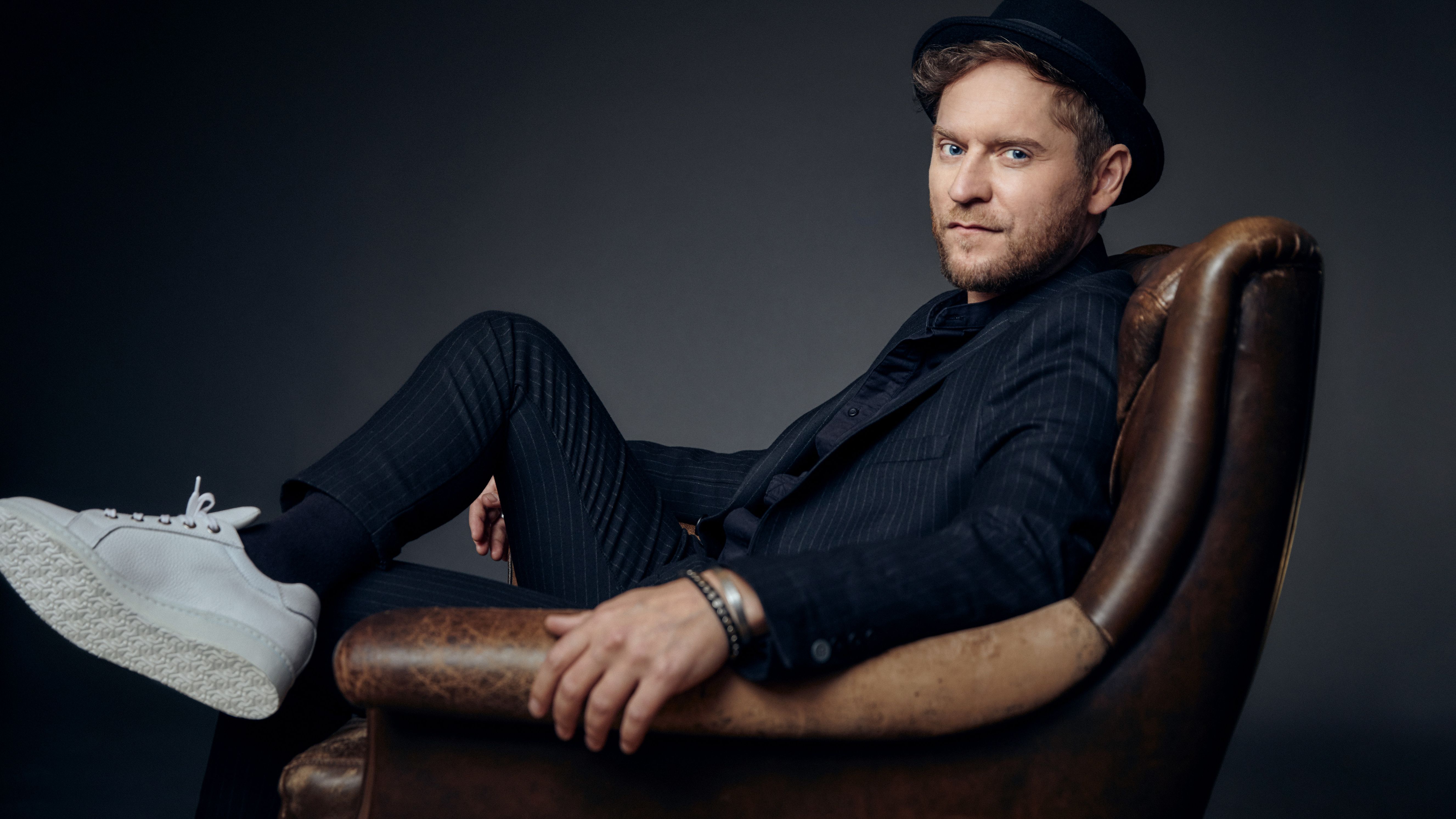 Johannes Oerding sitzt auf einem braunen Ledersessel. Er trägt einen schwarzen, gestreiften Anzug und einen Hut. 