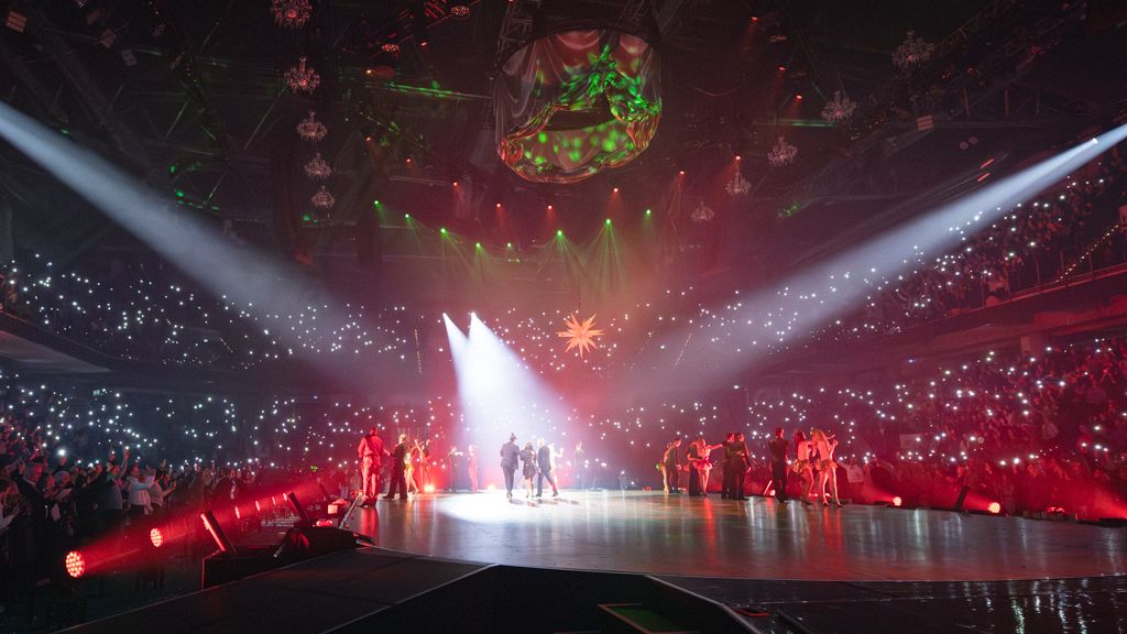 Auf der Bühne stehen mehrere Personen im Licht roter und weißer Scheinwerfer, während das Publikum auf den Rängen mit eingeschalteten Handylichtern eine funkelnde Atmosphäre erzeugt. Über der Bühne hängen Dekorationen und Lichtelemente, die Szene wirkt festlich.