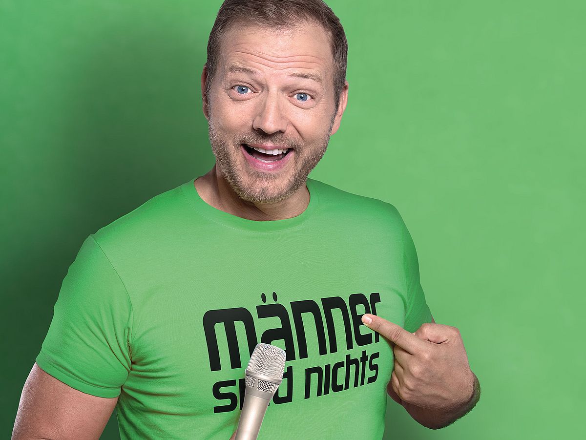 Mario Barth steht vor einem grünen Hintergrund. Er trägt ein T-Shirt in der identischen Farbe und hat ein Mikrofon in der Hand. 