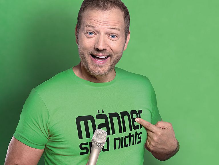 Mario Barth steht vor einem grünen Hintergrund. Er trägt ein T-Shirt in der identischen Farbe und hat ein Mikrofon in der Hand. 