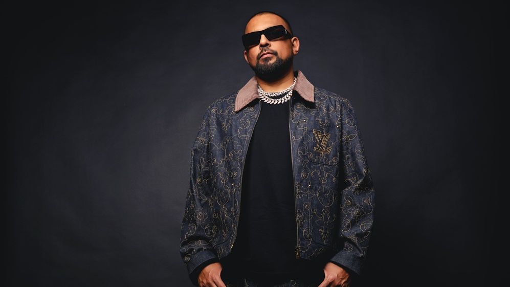 Sean Paul steht vor einem dunklen Hintergrund. Sean Paul trägt eine Sonnenbrille, eine zur Jeans passende Jeansjacke mit goldenen Verzierungen. Auf dem schwarzen Oberteil liegt eine silberne Kette. Er hat weiße Schuhe an. 