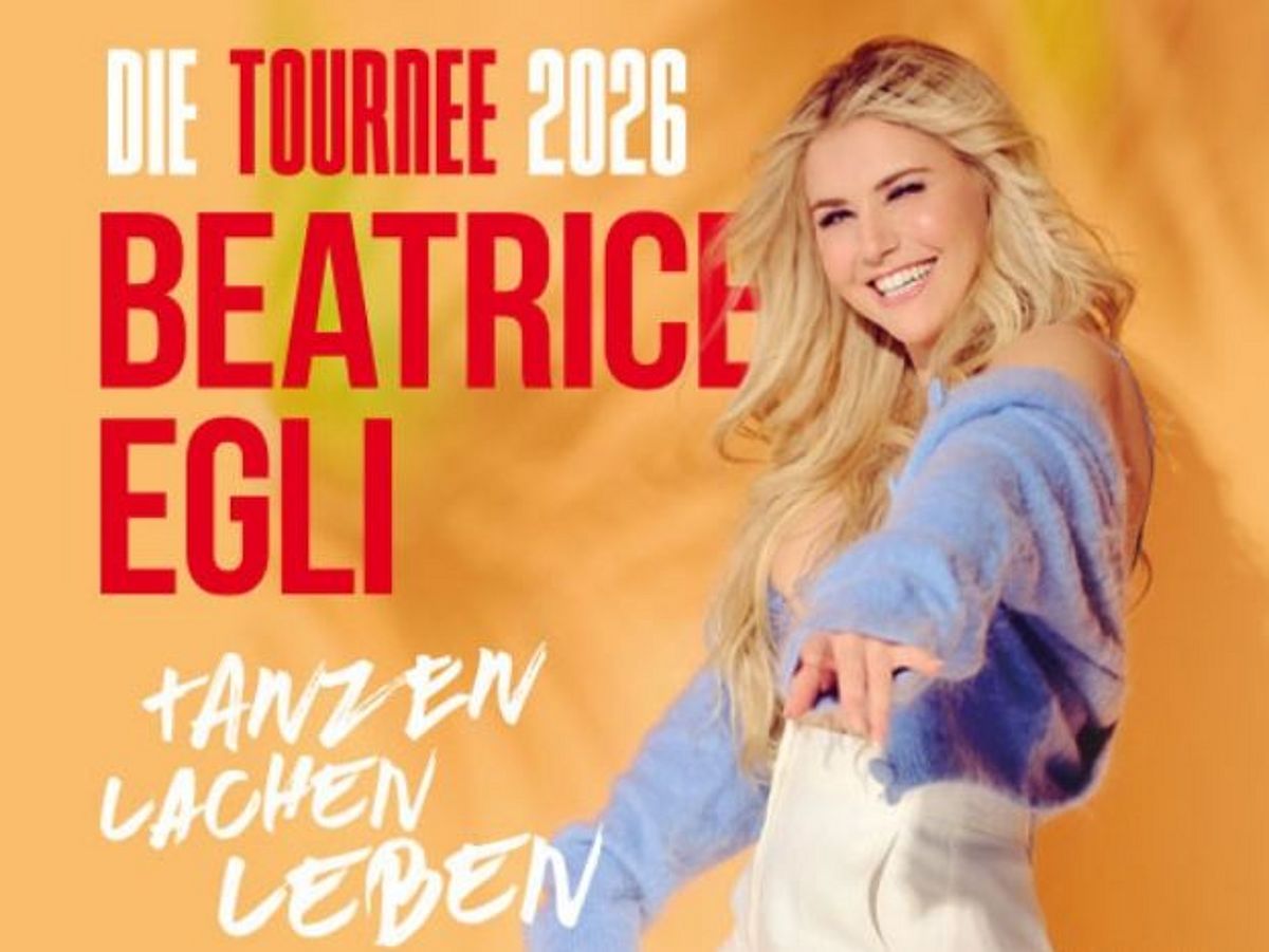 Beatrice Egli steht vor einem ockergelben Hintergrund. Sie trägt einen hellblaues Oberteil und eine weiße Hose.