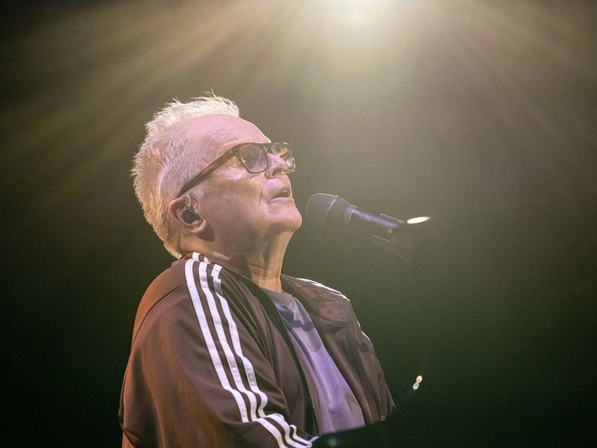 Herbert Grönemeyer sitzt in einem Scheinwerferlicht am Klavier und singt. Er trägt eine braune Adidas-Jacke und eine Brille. 