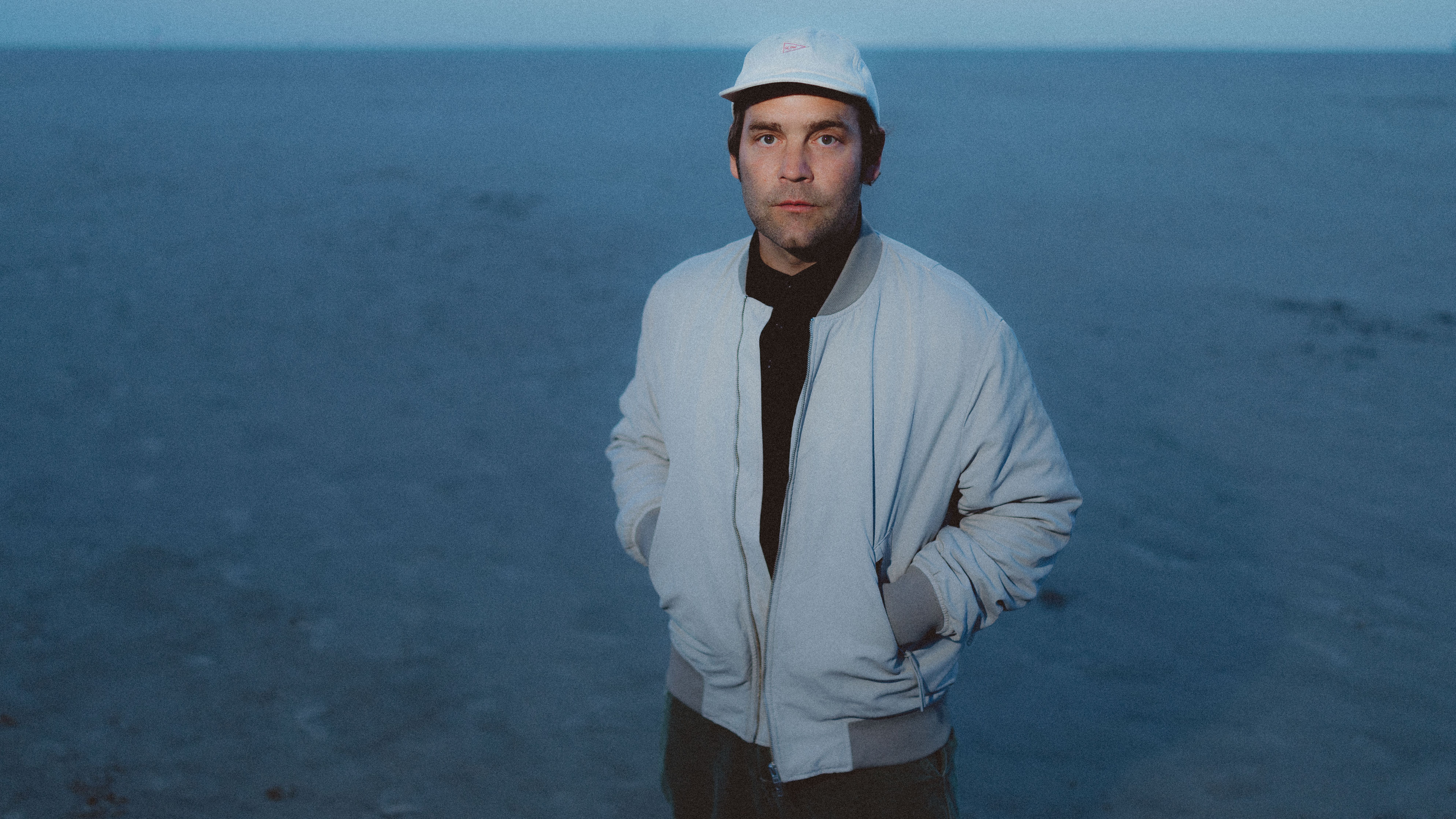 Bosse steht am Meer. Er trägt ein weißes Cap und eine beige Jacke. 