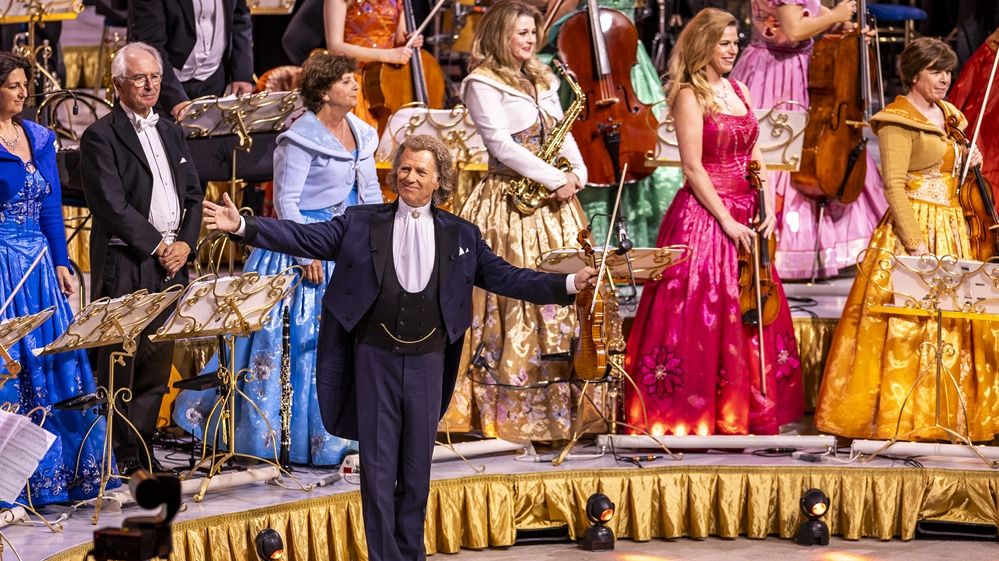 Eine Nahaufnahme von André Rieu, der auf der Bühne steht und seine Arme ausbreitet. Einige Orchestermitglieder sind im Hintergrund zu sehen, sie stehen und lächeln. André Rieu breitet die Arme aus. 