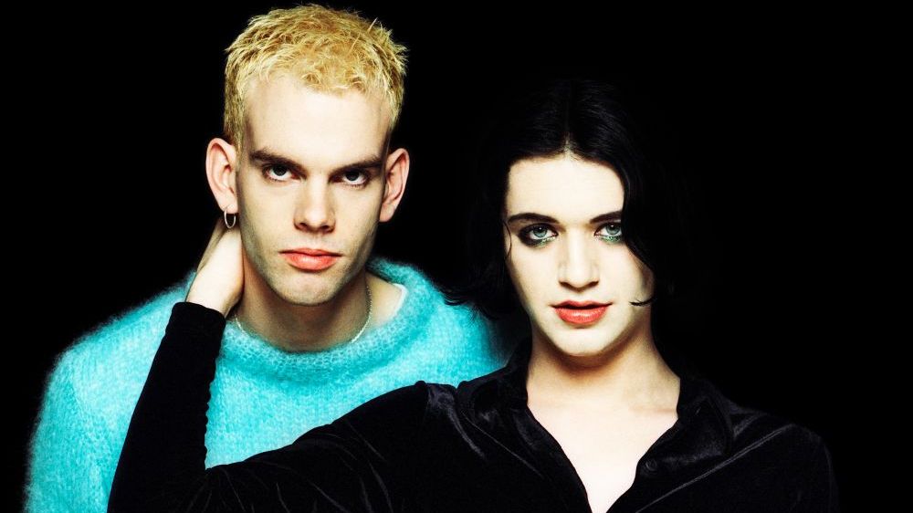 Pressefoto von Placebo mit Brian Molko im Vordergrund. Er trägt ein schwarzes Oberteil, seine Augen sind dunkel geschminkt. Seine rechte Hand ist am Hals eines anderen Bandmitglieds, der einen türkisen Wollpulli trägt. Er hat blondierte Haare und an einem Ohr einen Ohrring. 