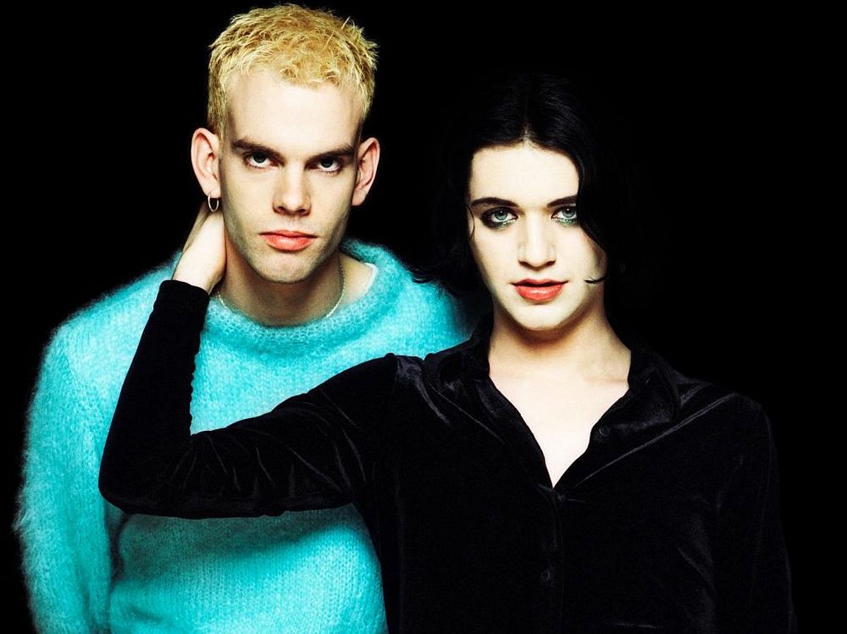 Pressefoto von Placebo mit Brian Molko im Vordergrund. Er trägt ein schwarzes Oberteil, seine Augen sind dunkel geschminkt. Seine rechte Hand ist am Hals eines anderen Bandmitglieds, der einen türkisen Wollpulli trägt. Er hat blondierte Haare und an einem Ohr einen Ohrring. 