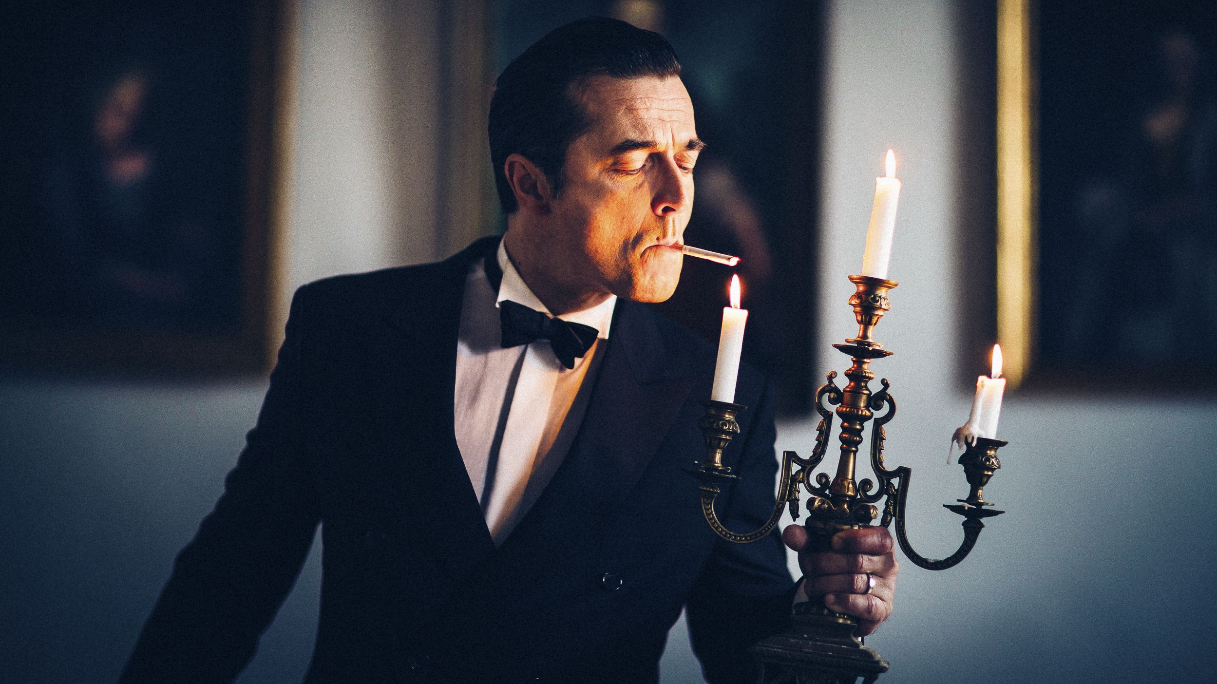 Ein Pressebild des FAlco-Darstellers, der einen Smoking trägt und an einem Kerzenständer eine Zigarette sich anzündet. 