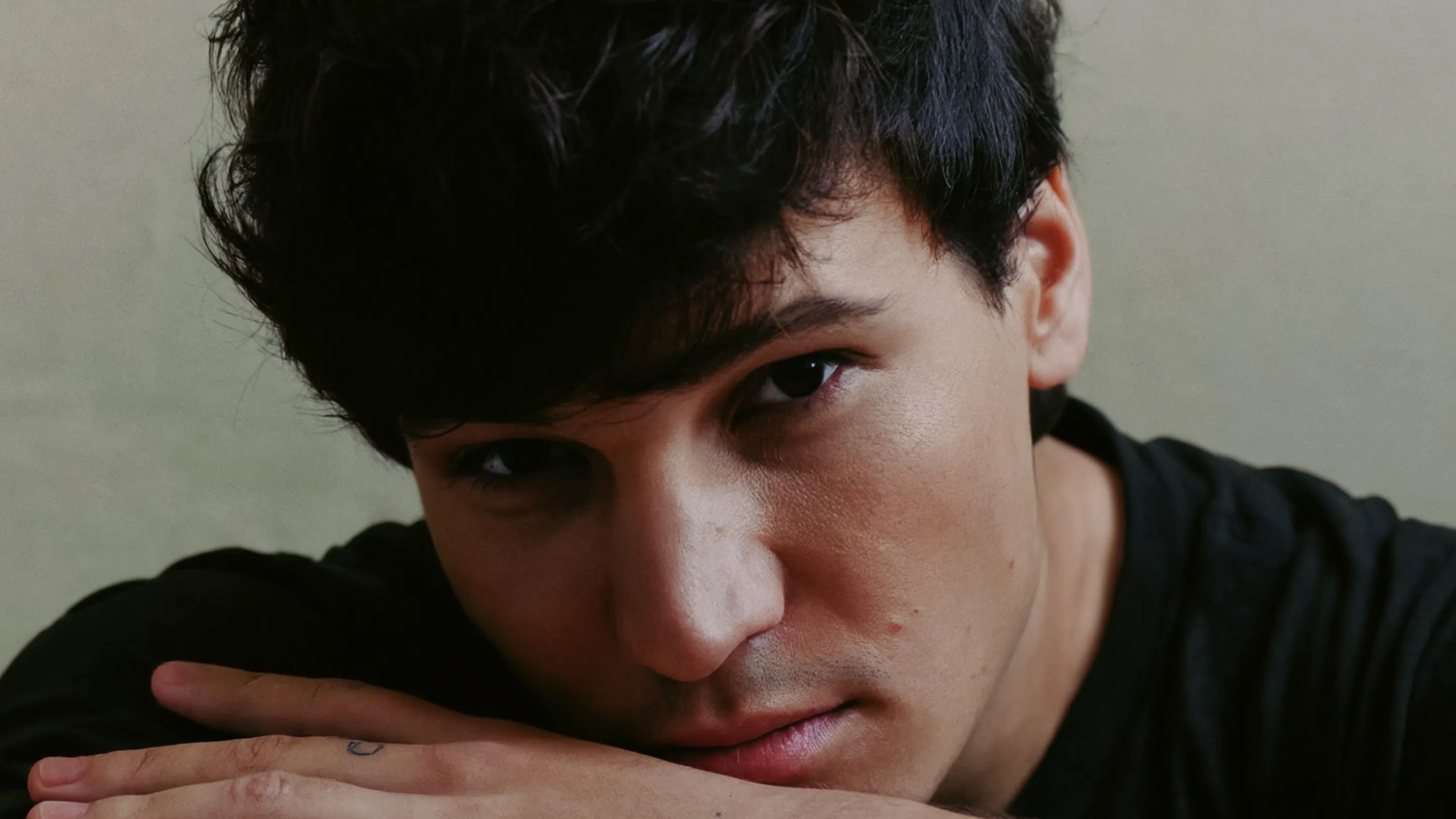 Pressebild von Wincent Weiss. Er steht vor einer weißen Wand, trägt dunkle Kleidung. Wincent Weiss schaut nach links und lächelt. Er hat kurze. lockige Haare. 