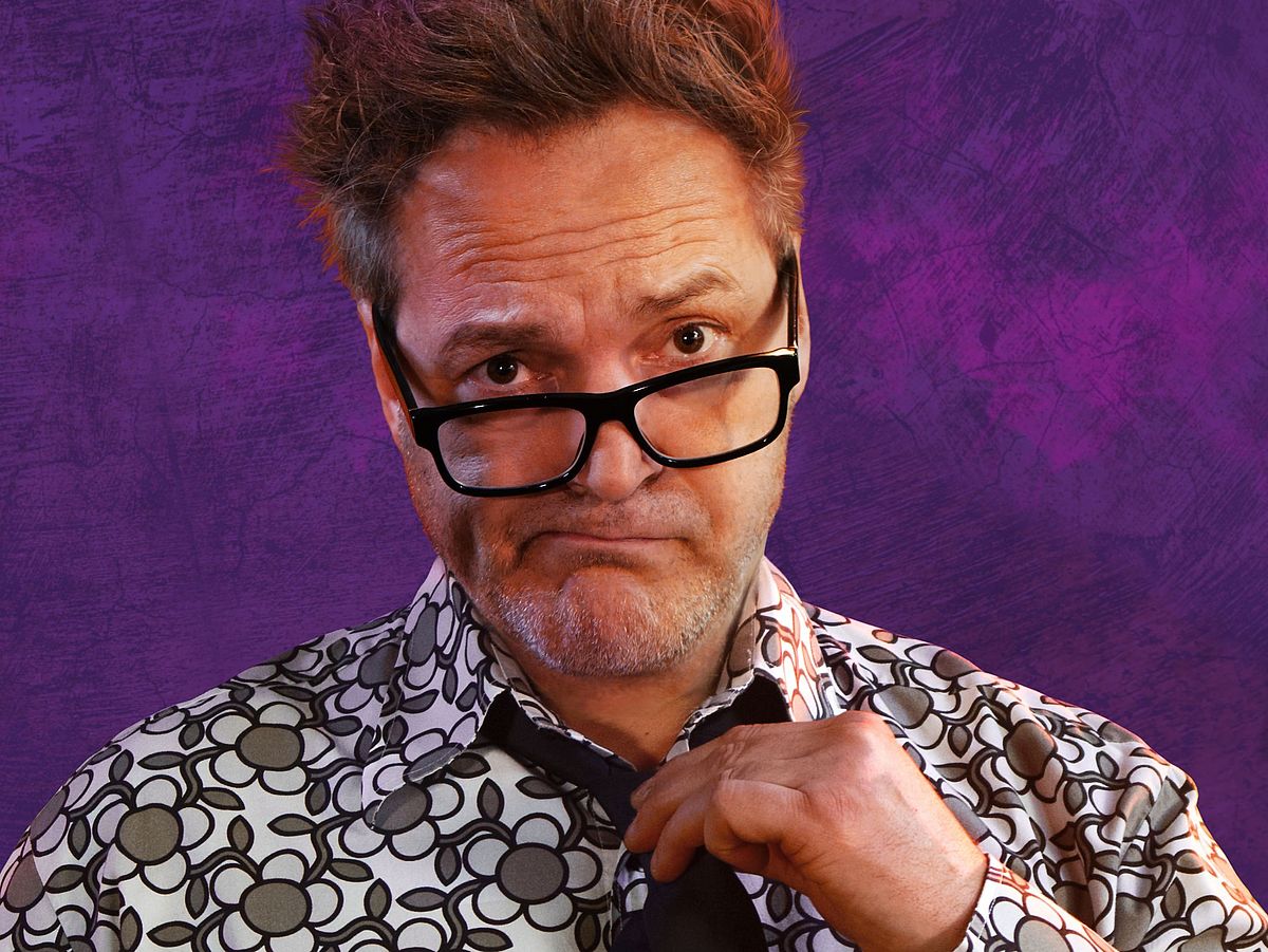 Paul Pnazer steht vor einem lila Hintergrund. Er trägt ein weiß-schwarz-grün-meliertes Hemd und eine schwarze Krawatte. 