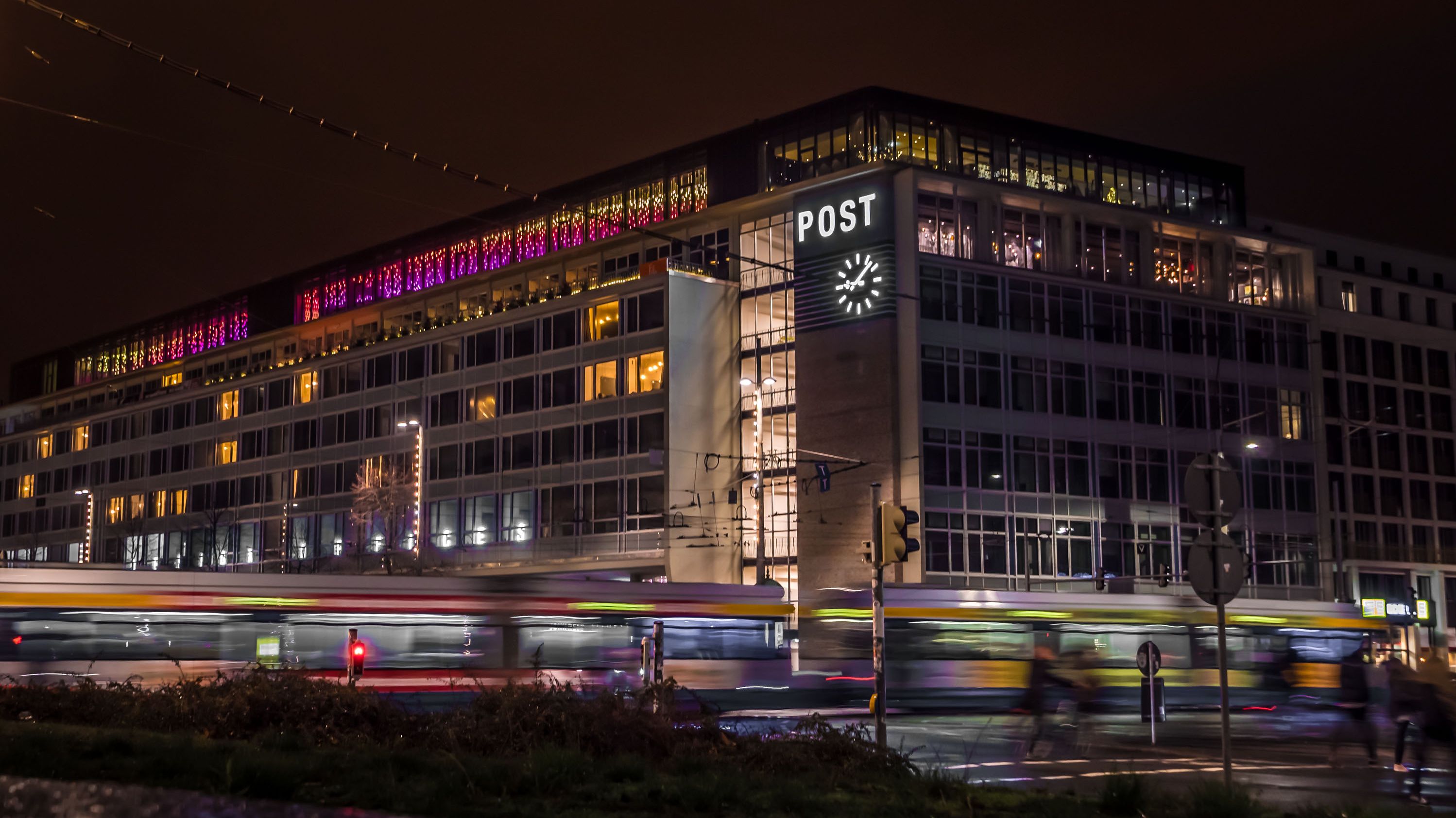 Blick auf das Hotel bei Nacht mit Beleuchtung. Vor dem Hotel fährt gerade eine Straßenbahn vorbei. Der Schriftzug Post mit einer Uhr ist auf dem Gebäude zu sehen. 