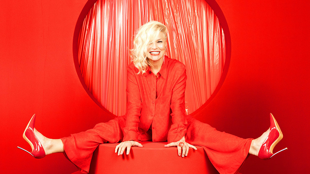 Ina Müller trägt rote Kleidung und sitzt auf einer roten Sitzmöglichkeit. Auch Boden und Hintergrund ist rot. 