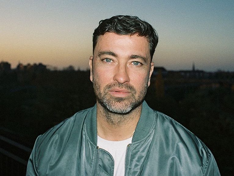 Marteria trägt eine grüne-blaue Bomberjacke und ein weißes T-Shirt. Er steht draußen, es ist Abenddämmerung, im Hintergrund ist eine Stadt zu sehen. Er hat einen Bart und grüne Augen. 