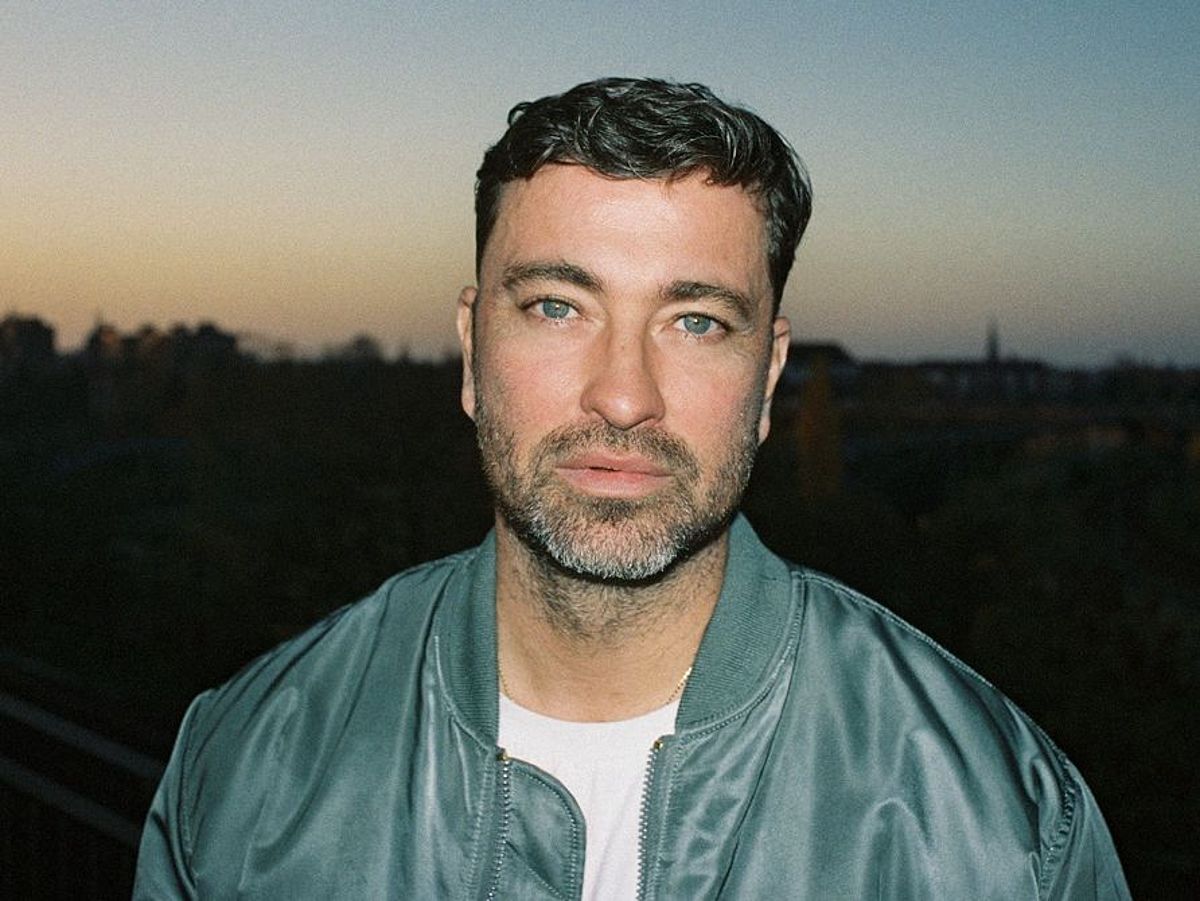 Marteria trägt eine grüne-blaue Bomberjacke und ein weißes T-Shirt. Er steht draußen, es ist Abenddämmerung, im Hintergrund ist eine Stadt zu sehen. Er hat einen Bart und grüne Augen. 