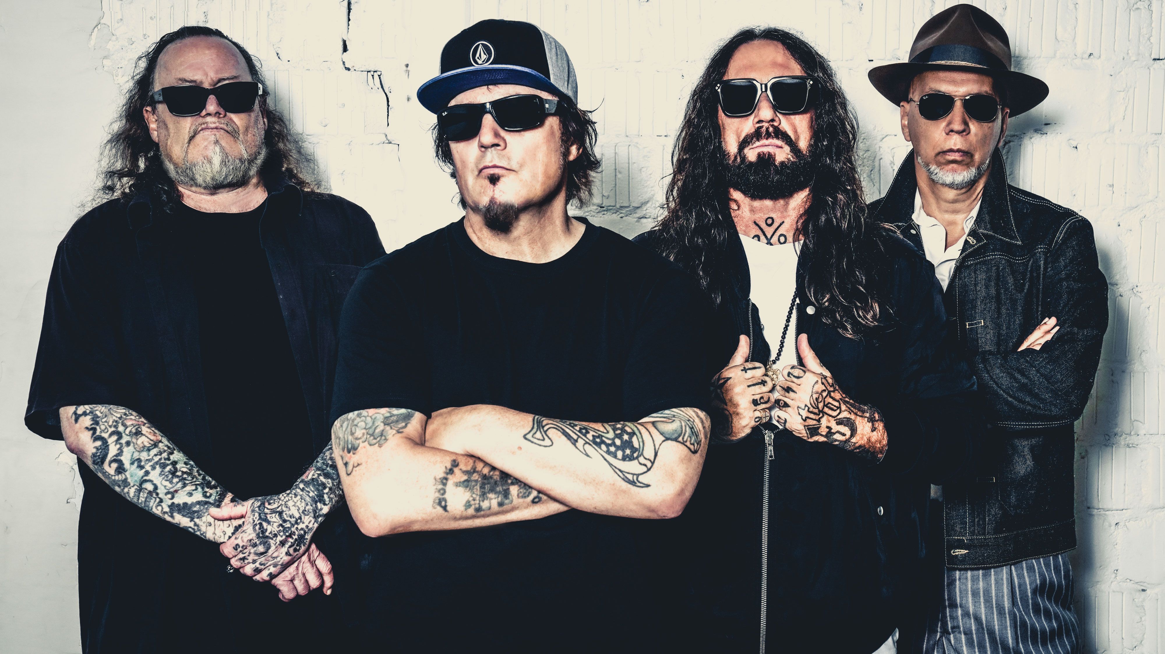 Die Band steht vor einer weißen Wand. Sie haben alle Tatoos, tragen schwarze Kleidung und Sonnenbrillen. 