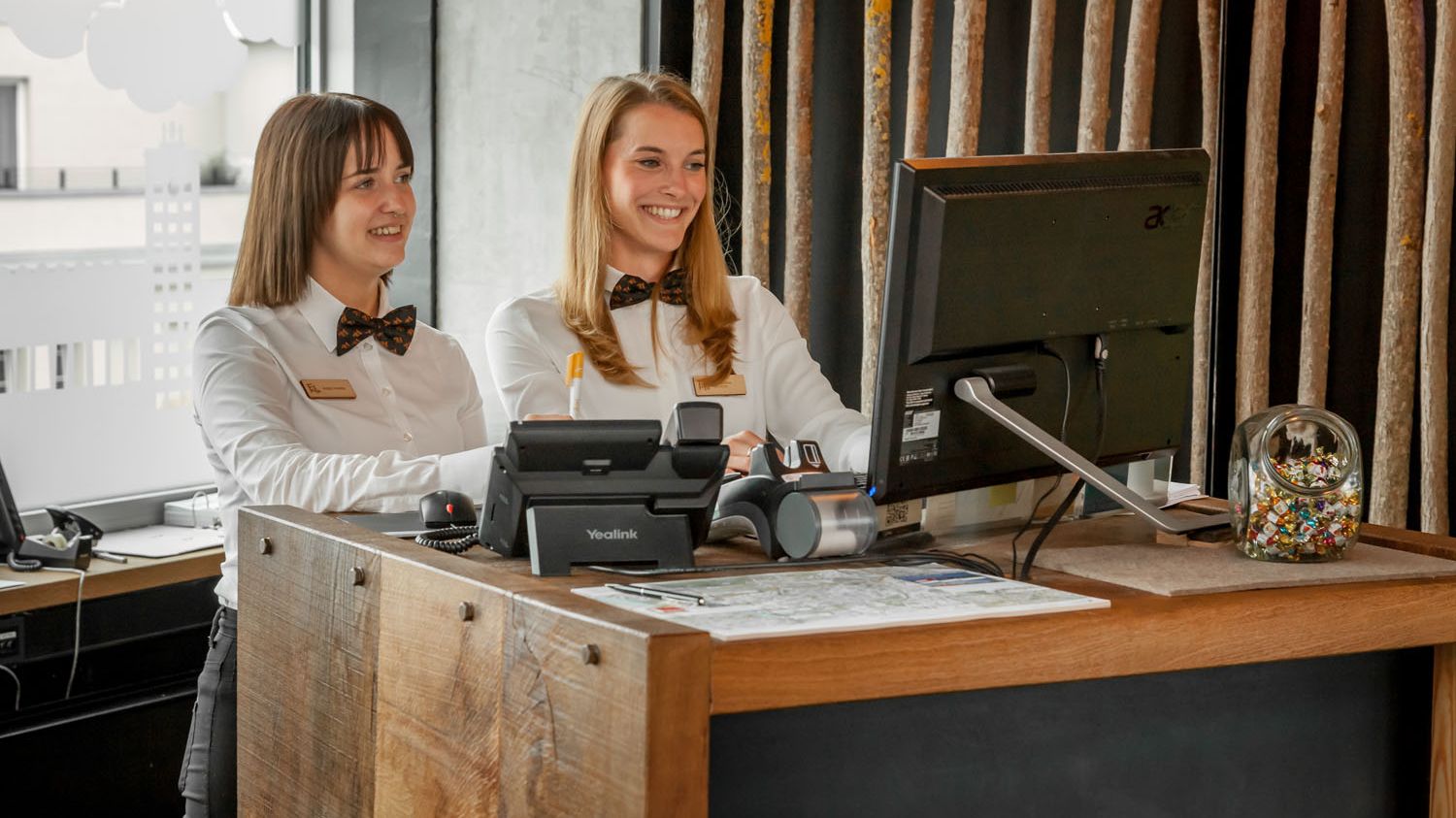 An der Hoterezeption stehen zwei Hotelmitarbeiterinnen und blicken auf einen Computer. Sie lächeln und tragen ein weißes Hemd mit Fliege. 