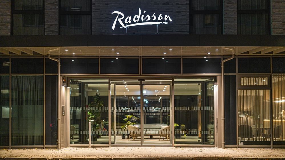 Außenansicht des Hotels bei Nacht. Der Eingangsbereich mit Glastüren ist beleuchtet.  