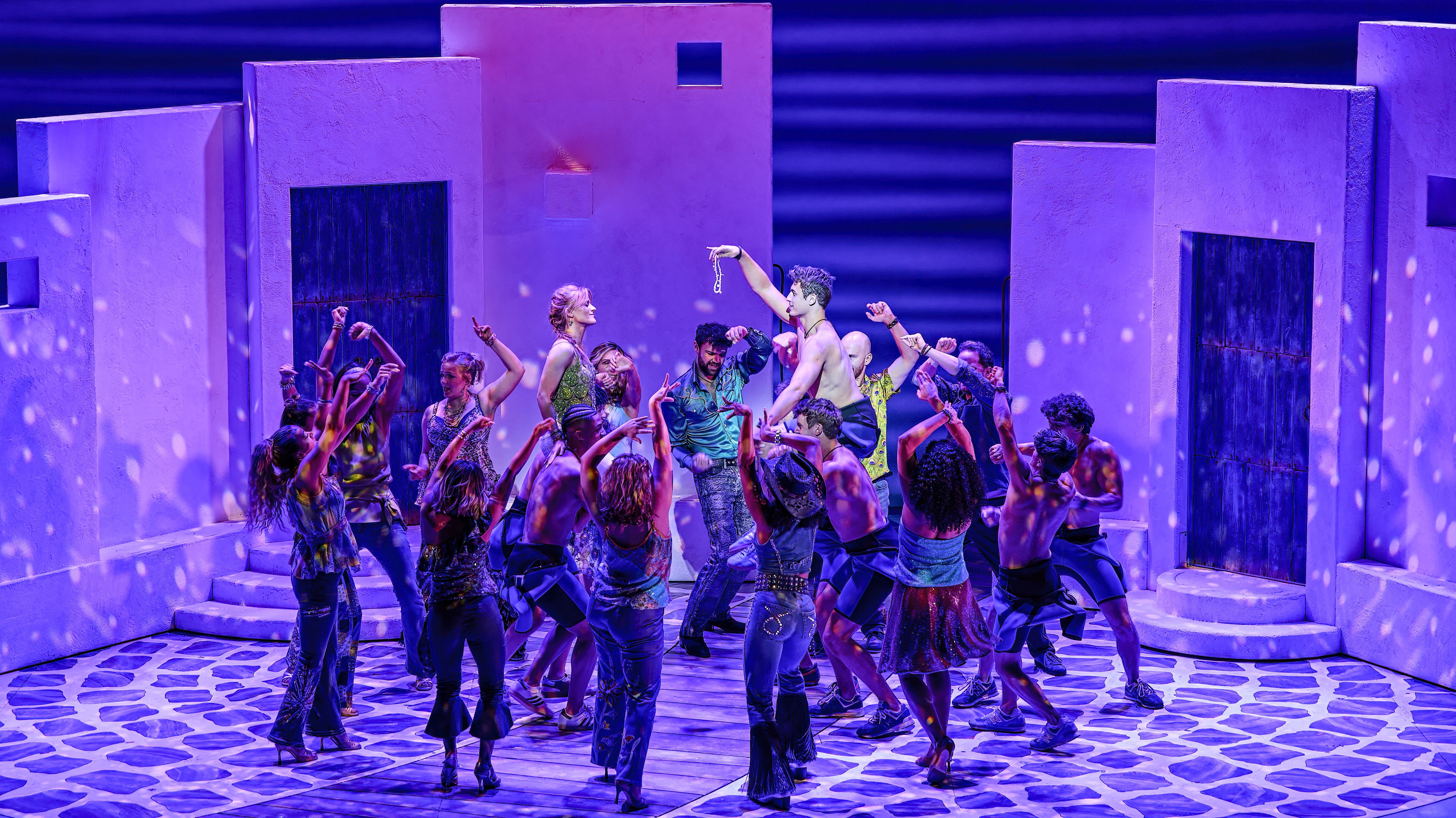 In der lila erleuchteten Kulisse eines Innenhofs trägt eine tanzende Gruppe von Männern und Frauen eine junge Frau und einen jungen Mann auf den Schultern. Der junge Mann hält der Frau eine Kette vor Augen.