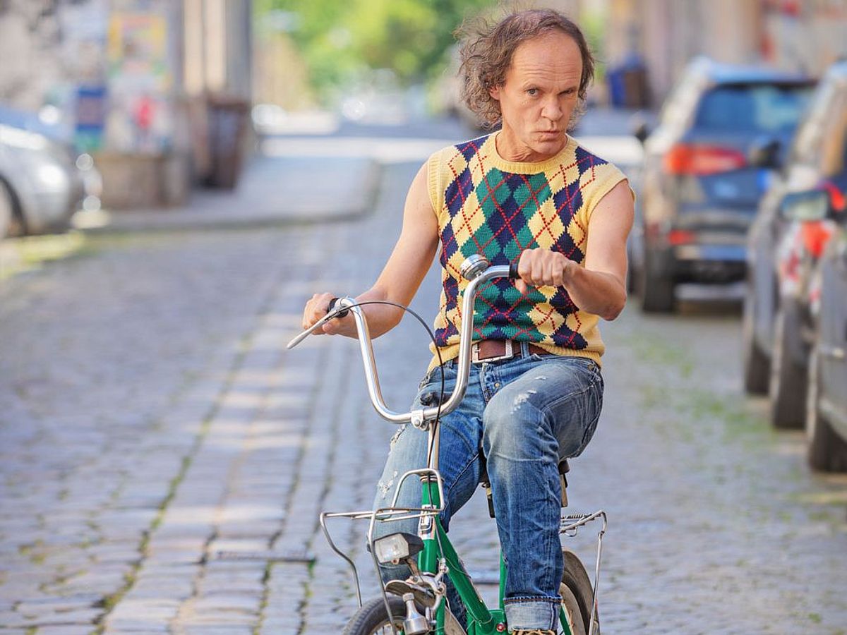 Olaf Schubert sitzt auf einem grünen Fahrrad, er trägt einen gelb-karierten Pullunder und eine Jeans. 