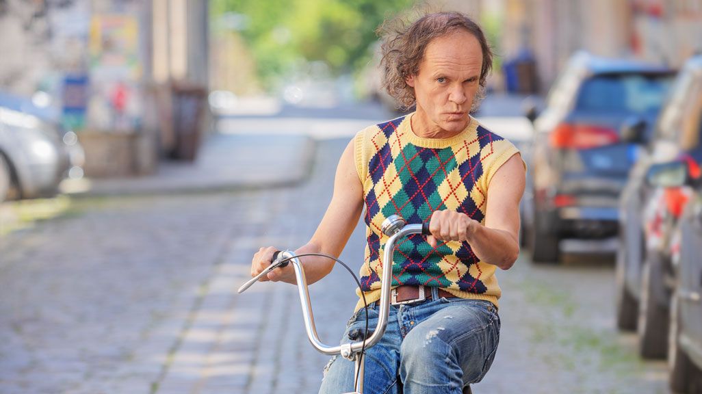 Olaf Schubert sitzt auf einem grünen Fahrrad, er trägt einen gelb-karierten Pullunder und eine Jeans. 