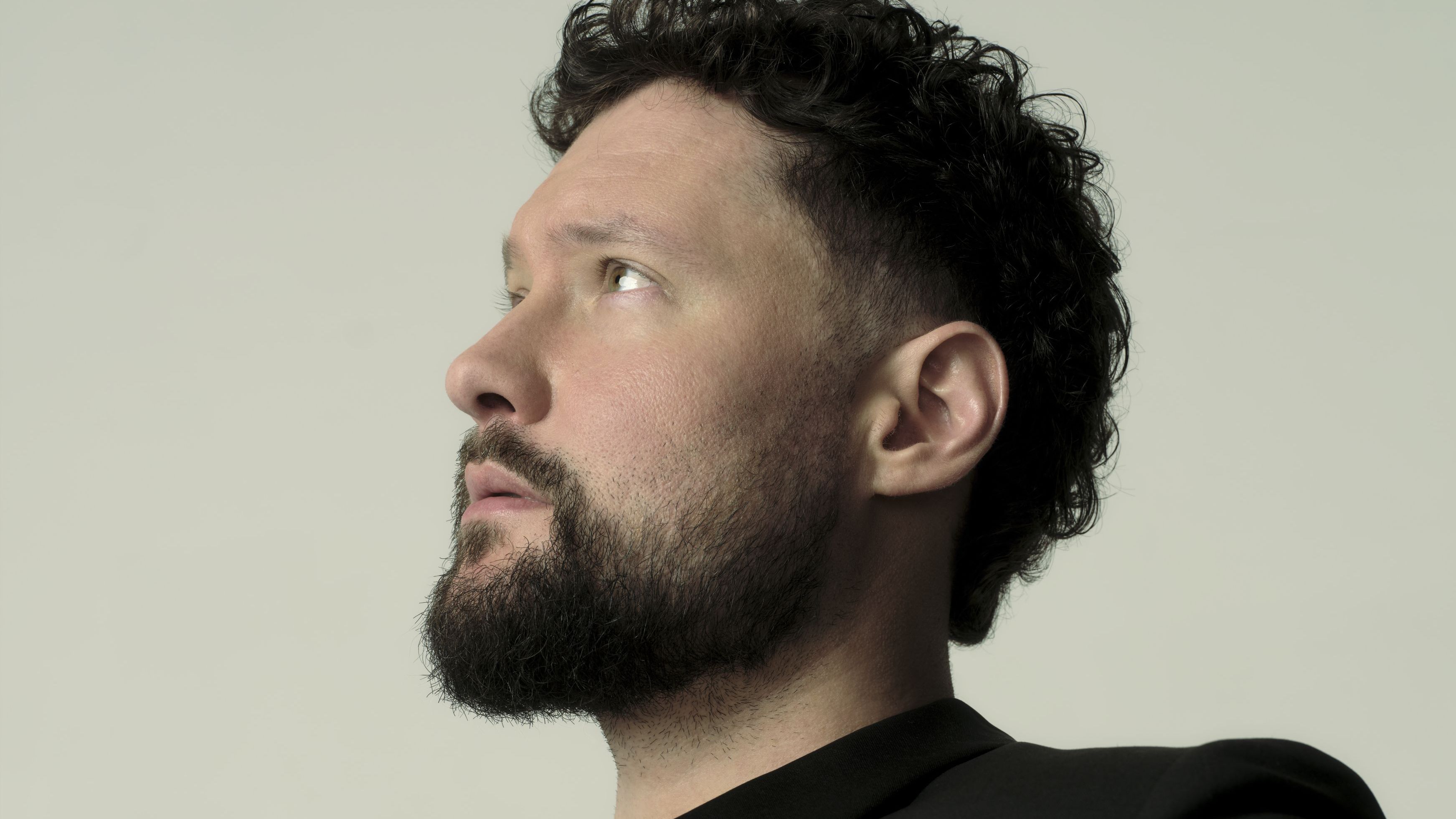 Calum Scott sitzt seitwärts auf einem Stuhl. Er trägt ein schwarzes Jackett, an diesem ist eine goldene Kette mit Anhänger. 