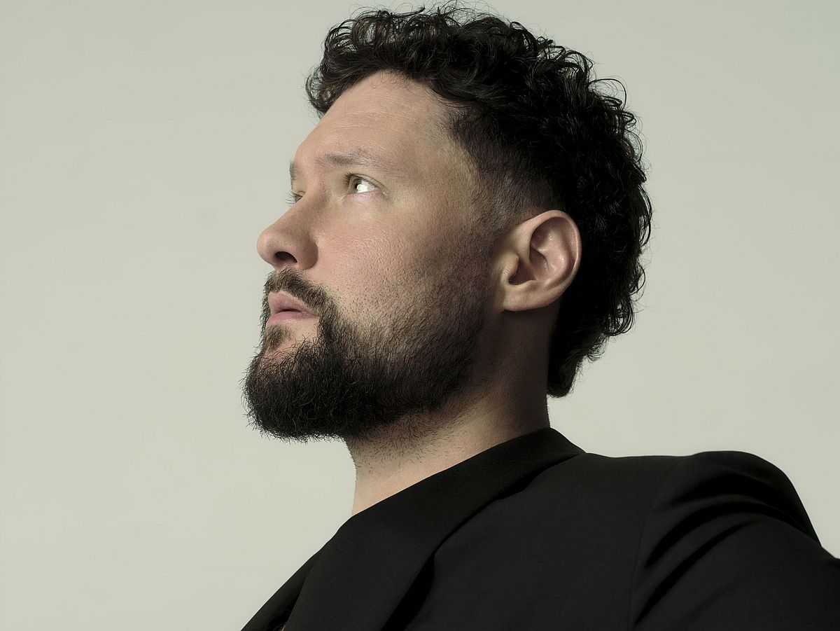 Calum Scott sitzt seitwärts auf einem Stuhl. Er trägt ein schwarzes Jackett, an diesem ist eine goldene Kette mit Anhänger. 