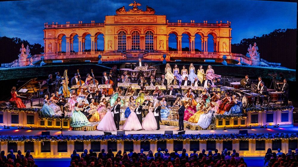 Ein Bild von einem Konzert von André Rieu mit großem Orchester. Auf der Bühne ist das Orchester und Sängerinnen und Sänger zu sehen. André Rieu steht seitlich mit einer Geige in der Hand. Alle Männer tragen einen Smoking, die Frauen Ballkleider. 