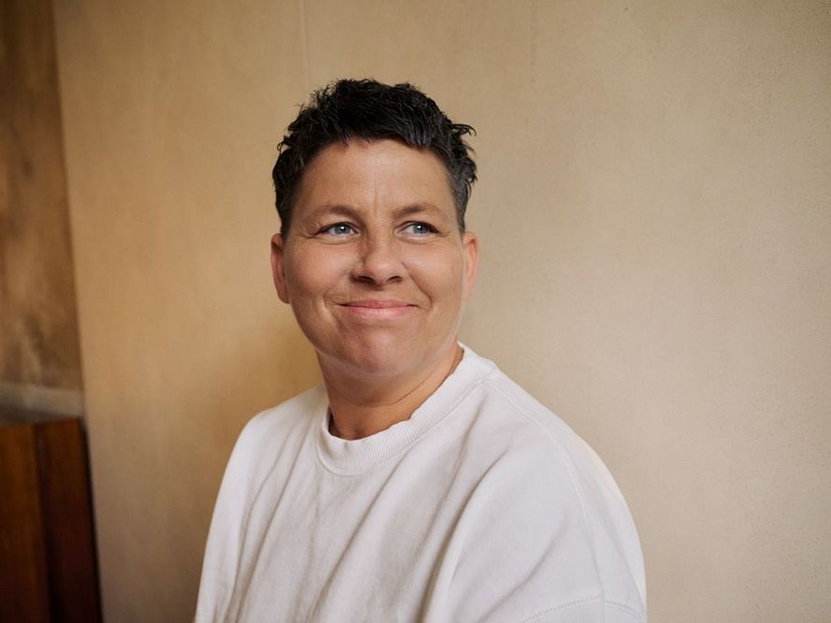 Kerstin Ott steht vor einer beigen Wand und schaut nach rechts. Kerstin Ott lächelt und trägt einen beigen Pullover. 