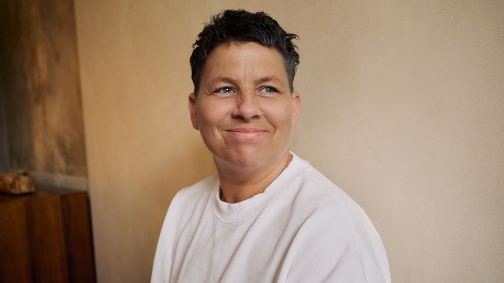 Kerstin Ott steht vor einer beigen Wand und schaut nach rechts. Kerstin Ott lächelt und trägt einen beigen Pullover. 