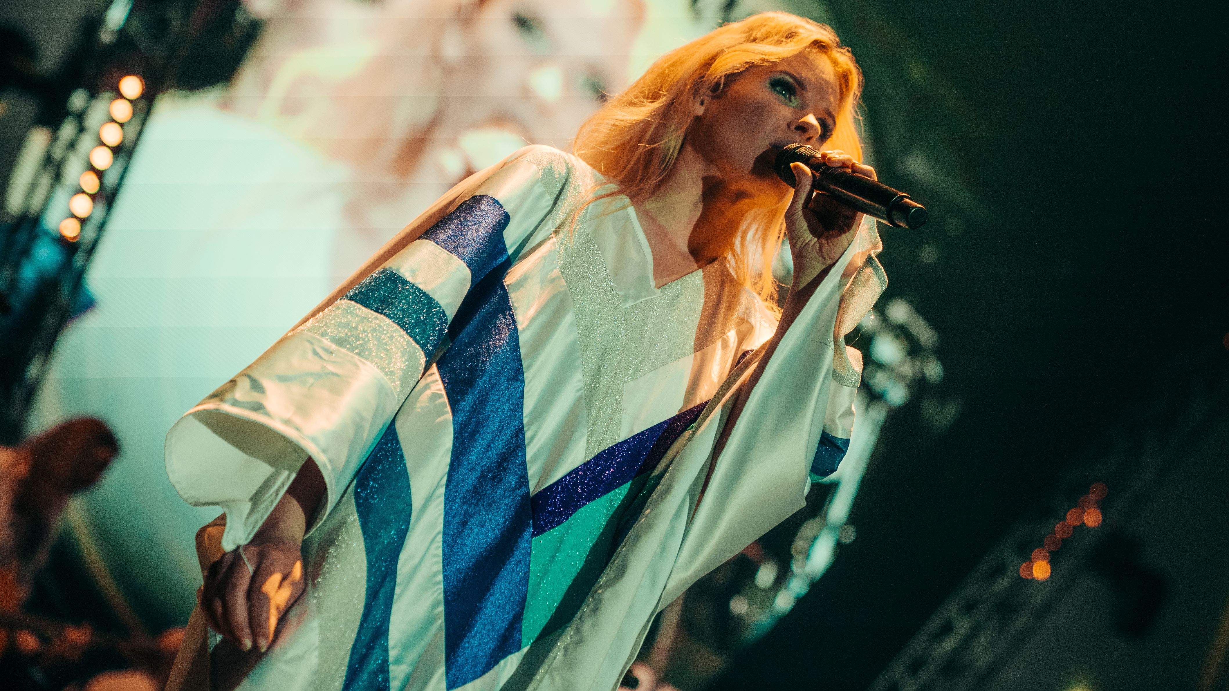 Eine Sängerin mit blonden Haaren ist zu sehen, die ein blau-weißes Cape trägt. 