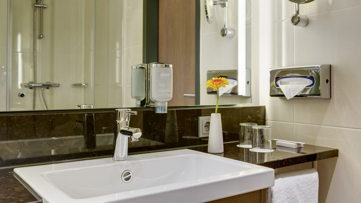 Bild eines Badezimmers mit dunklen Marmorarmaturen. Man sieht ein weißes Waschbecken, im Spiegel sieht man die Dusche. 