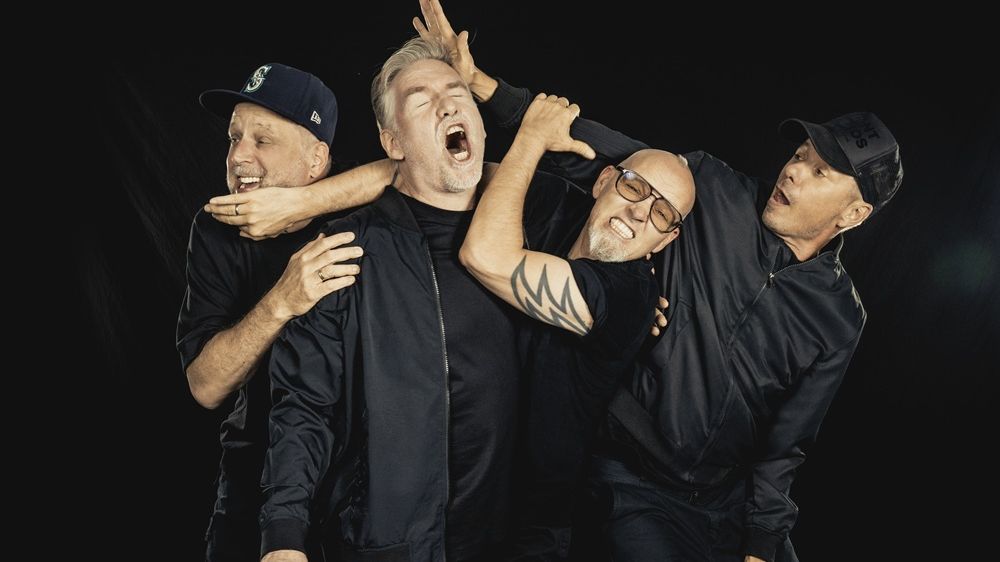 Die Bandmitgleider tragen alle schwarze Kleidung. Sie posieren humorvoll vor schwarzem Hintergrund. Sie stehen eng beieinander und tun so als ob sie sich gegenseitig schubsen oder umarmen würden. 