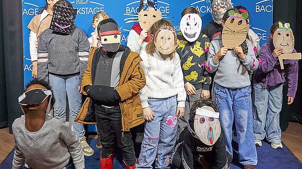 15 Kinder stehen in einer Gruppe vor einer blauen Fotowand, auf der viele Logos des BACKSTAGE CLUBS zu sehen sind. Die Kinder tragen selbst gebastelte Masken aus Pappe. Manche Kinder knien, die meisten stehen.  