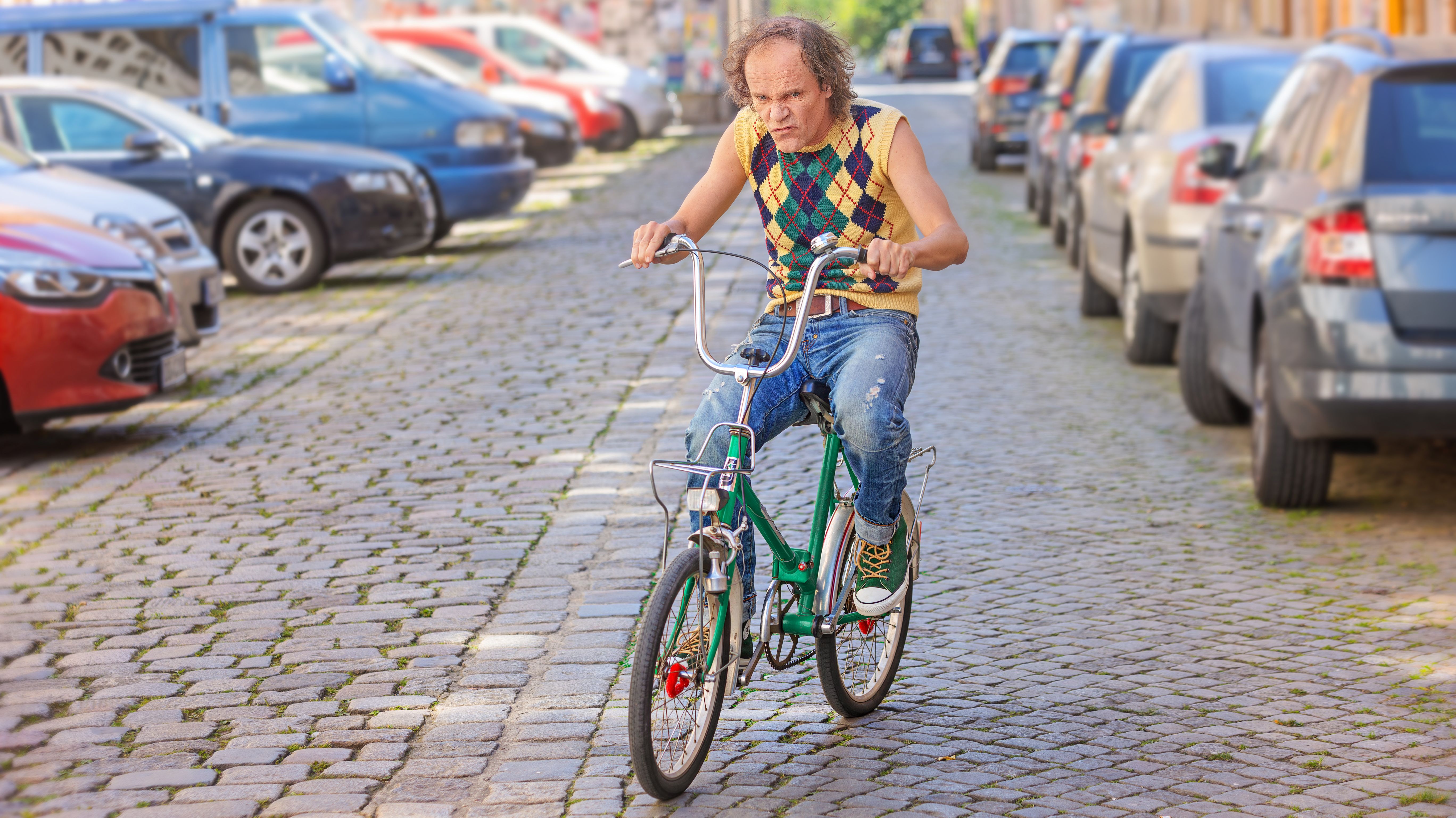 Olaf Schubert fährt mit einem kleinen grünen Fahrrad über eine Kopfsteinpflasterstraße, die von parkenden Autos gesäumt ist.