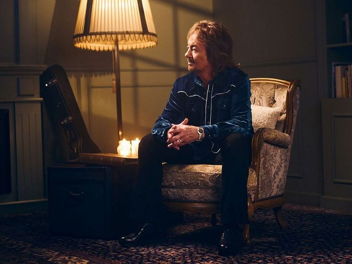 Chris Norman sitzt in einem Sessel in einem Wohnzimmer, neben ihm ist eine Stehlampe an. Im Hintergrund ist ein Bücherregal zu sehen. 