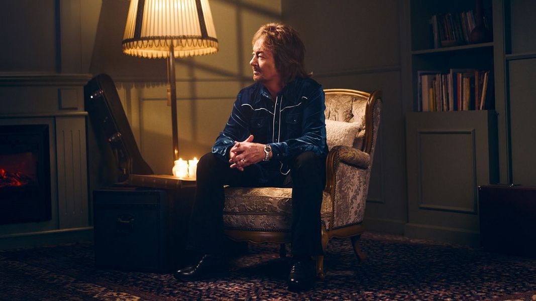 Chris Norman sitzt in einem Sessel in einem Wohnzimmer, neben ihm ist eine Stehlampe an. Im Hintergrund ist ein Bücherregal zu sehen. 