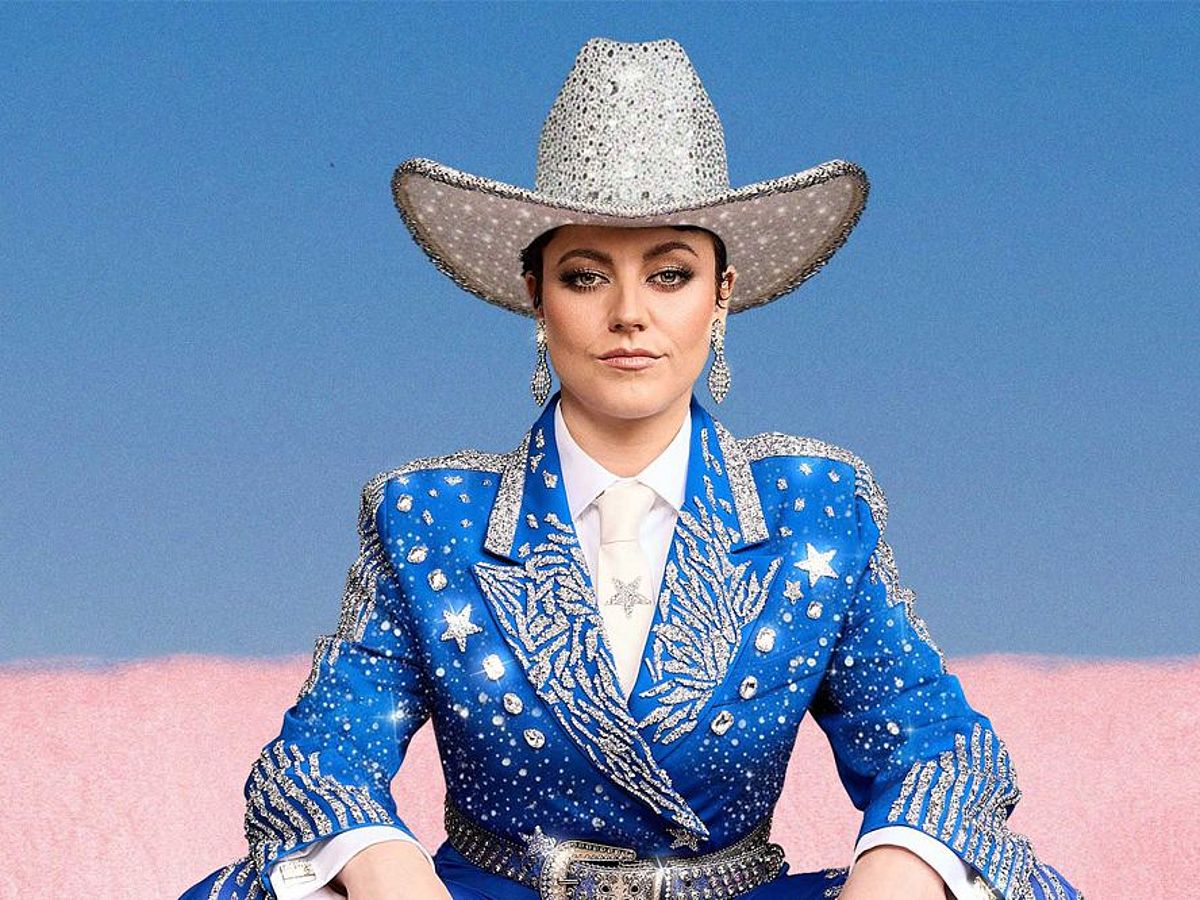 Tahnee sitzt in einem blau-glitzernden Anzug un einem weiß-silbrigen Cowboy-Hut vor einem rosa-hellblauen Hintergrund. sie trägt glitzernde Ohrringe. 