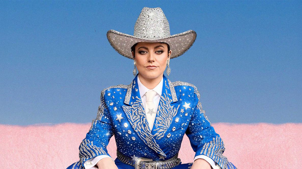 Tahnee sitzt in einem blau-glitzernden Anzug un einem weiß-silbrigen Cowboy-Hut vor einem rosa-hellblauen Hintergrund. sie trägt glitzernde Ohrringe. 