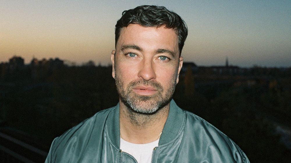 Marteria trägt eine grüne-blaue Bomberjacke und ein weißes T-Shirt. Er steht draußen, es ist Abenddämmerung, im Hintergrund ist eine Stadt zu sehen. Er hat einen Bart und grüne Augen. 