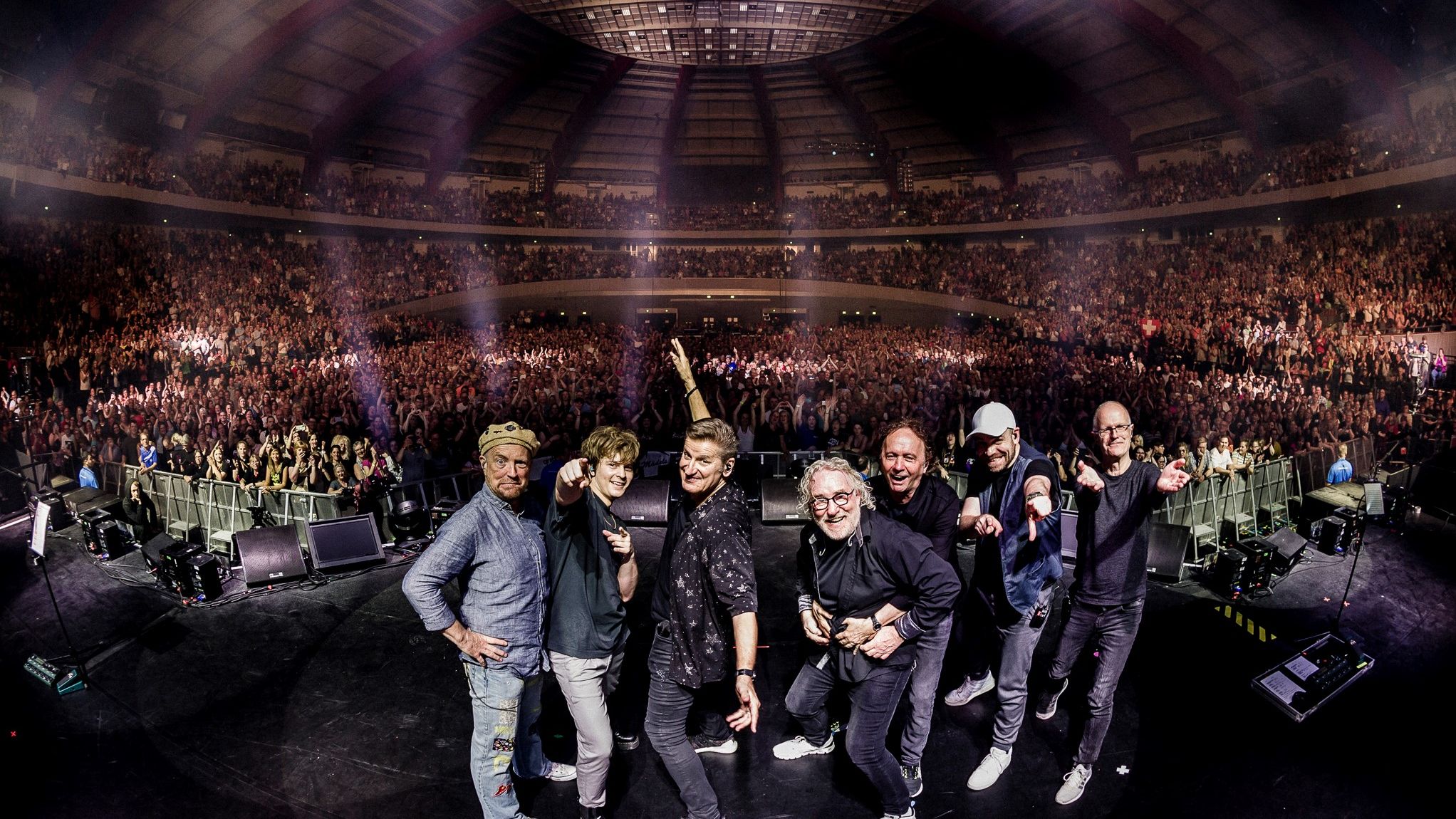 Die Band steht auf einer Bühne mit dem Rücken zum Publikum. Im Hintergrund ist das Publikum zu sehen