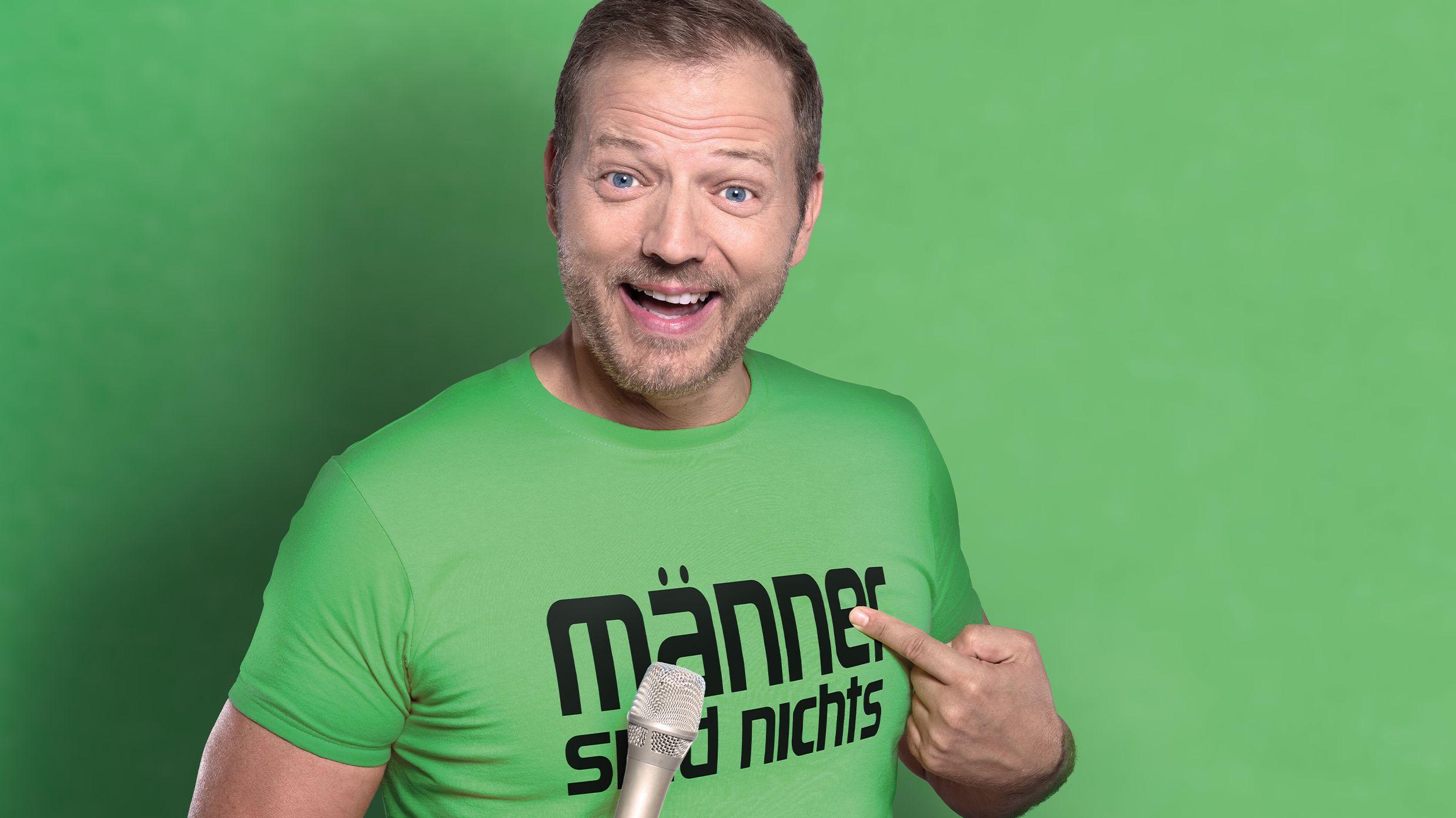 Mario Barth steht vor einem grünen Hintergrund. Er trägt ein T-Shirt in der identischen Farbe und hat ein Mikrofon in der Hand.