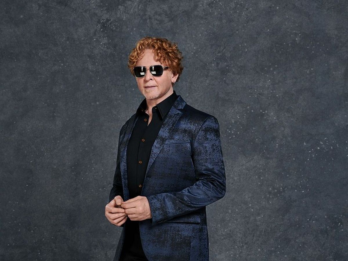 Mick Hucknall steht vor einem grauen Hintergrund. Er trägt ein dunkelblaues Jacket und ein schwarzes Hemd. Er hat eine dunkle, reflektierende Sonnenbrille auf. 