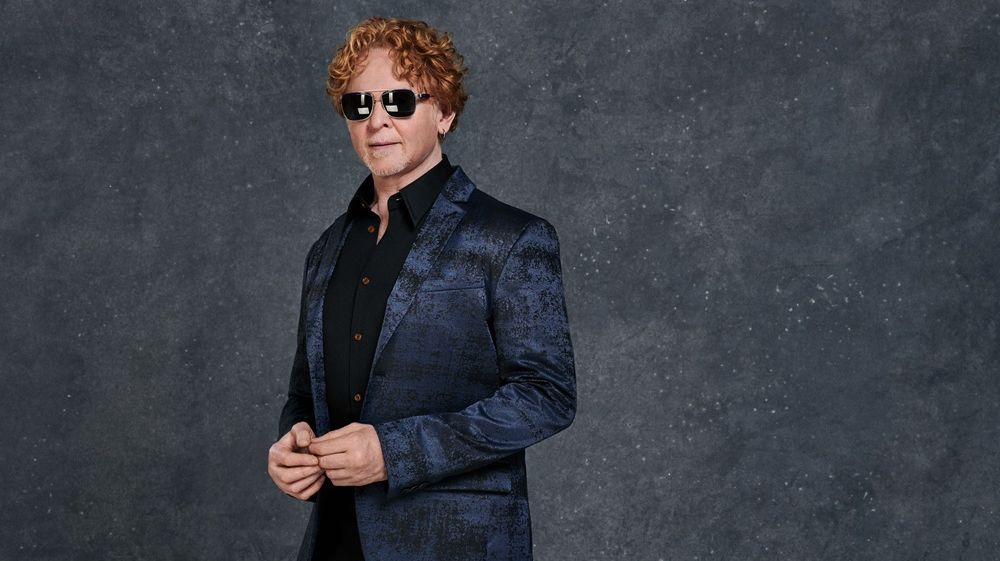 Mick Hucknall steht vor einem grauen Hintergrund. Er trägt ein dunkelblaues Jacket und ein schwarzes Hemd. Er hat eine dunkle, reflektierende Sonnenbrille auf. 